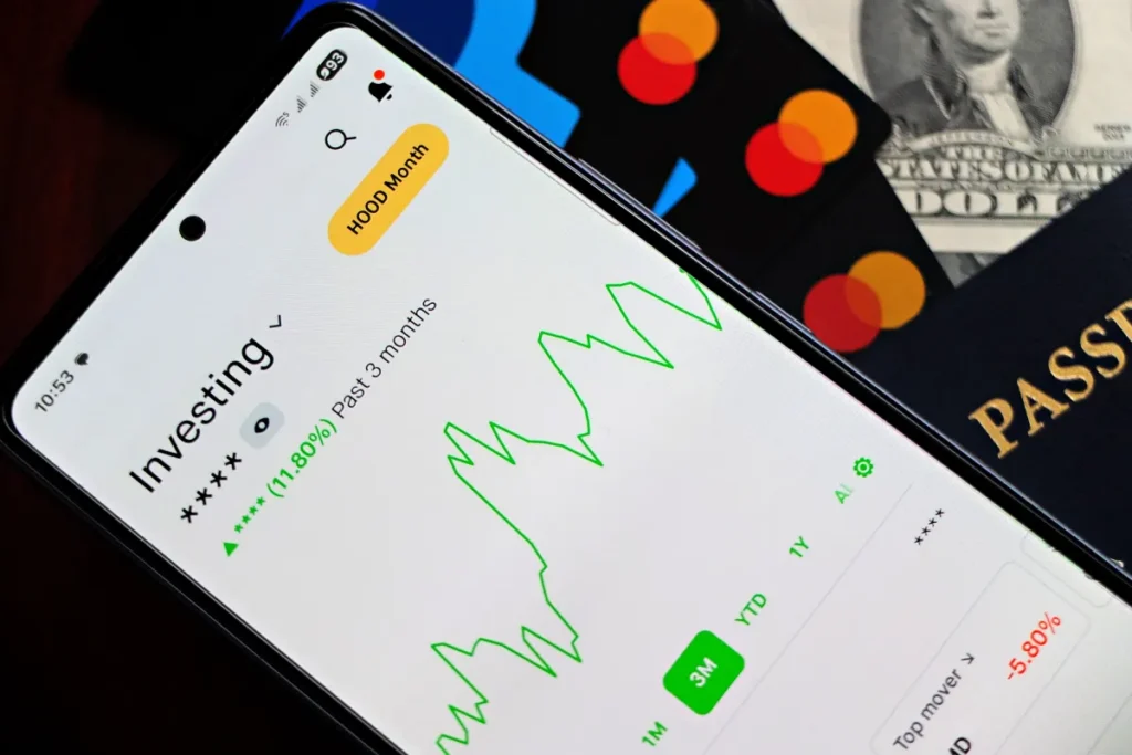 Are you building your portfolio one headline at a time? - A smartphone showing an investment app with green growth indicators, surrounded by credit cards, US dollars, and a passport.